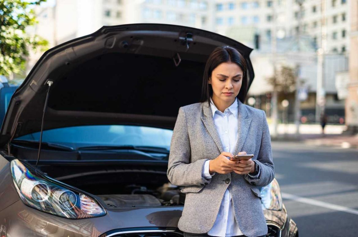 Femme inquiète utilisant son smartphone devant sa voiture en panne au capot ouvert, recherchant une assurance temporaire.